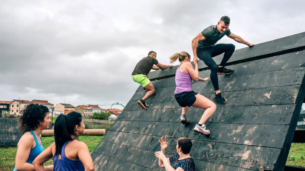 Group of people participating in an obstacle course, helping each other climb a sloping wall outdoors.